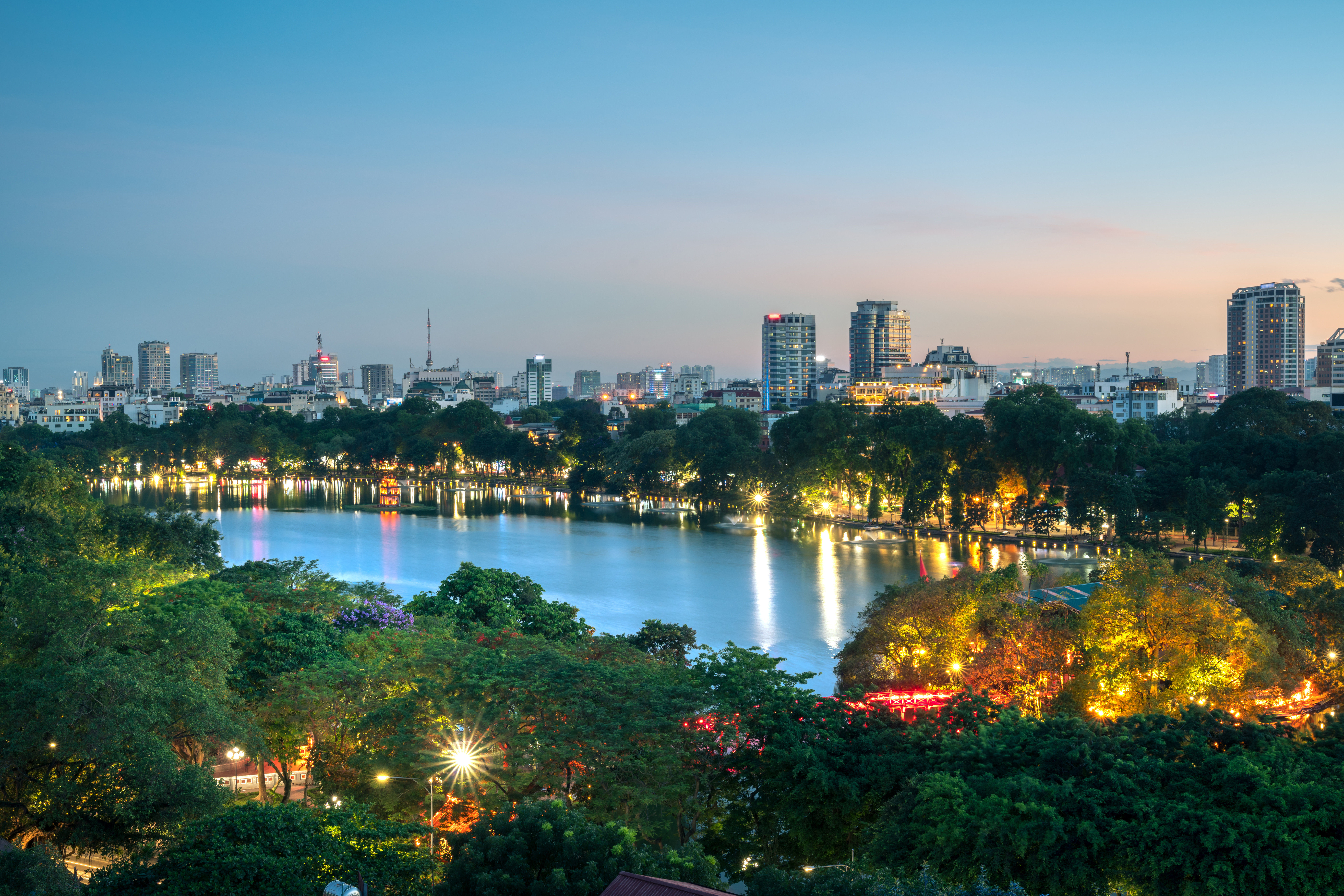 Skyline av Hanoi, Vietnam i skumringen med Hoan Kiem-sjøen i forgrunnen, omgitt av opplyste bybygninger og livlig grøntområder