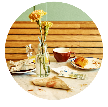 Frokostbord med blomster i en vase, en kopp, croissant på en tallerken og en fem-euroseddel.