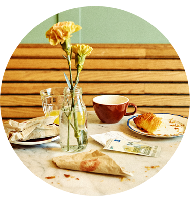 Blomster i en vase ved siden av en kaffekopp, croissant og euroseddel på kafébord.