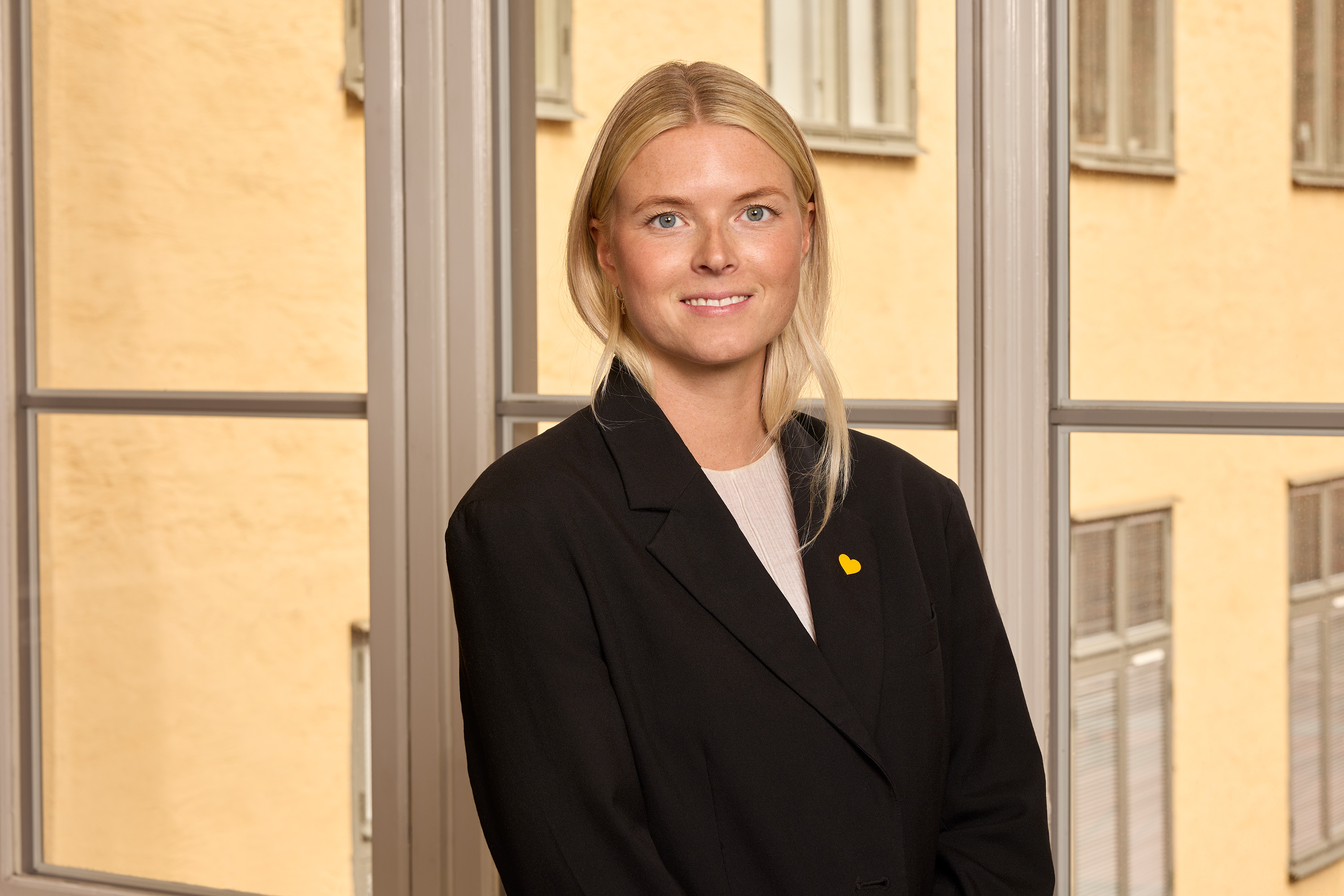Businesswoman in black jacket stands in front of a large window overlooking yellow building.