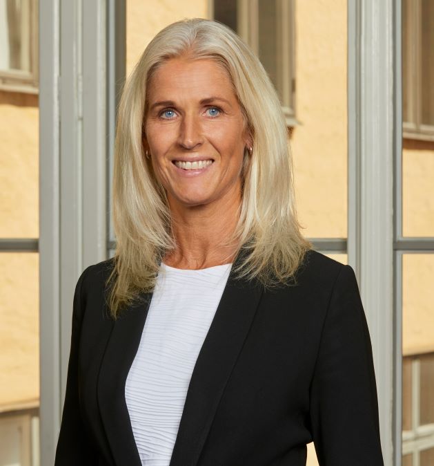 A woman with blonde hair in a black jacket stands in front of a window.