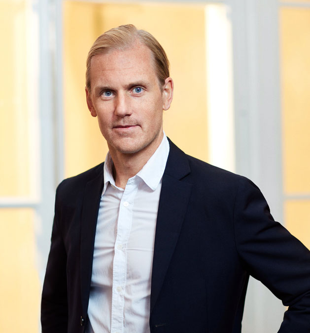 Man in black jacket and white shirt stands in front of bright windows.