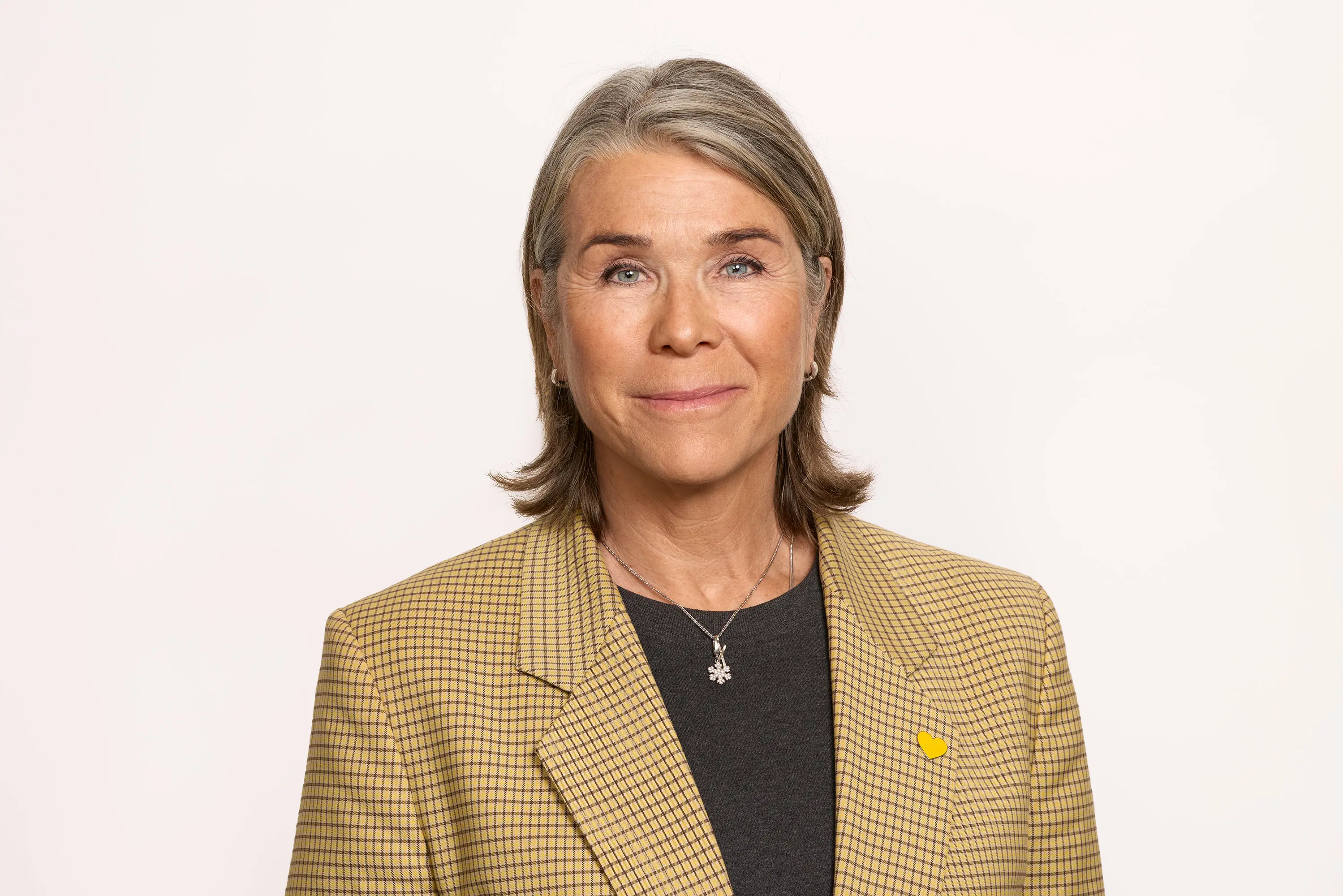 Kvinne i rutete blazer med grå genser, smilende foran en lys bakgrunn.