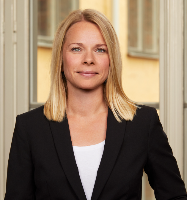 Woman in black blazer with long blonde hair stands in front of a window.