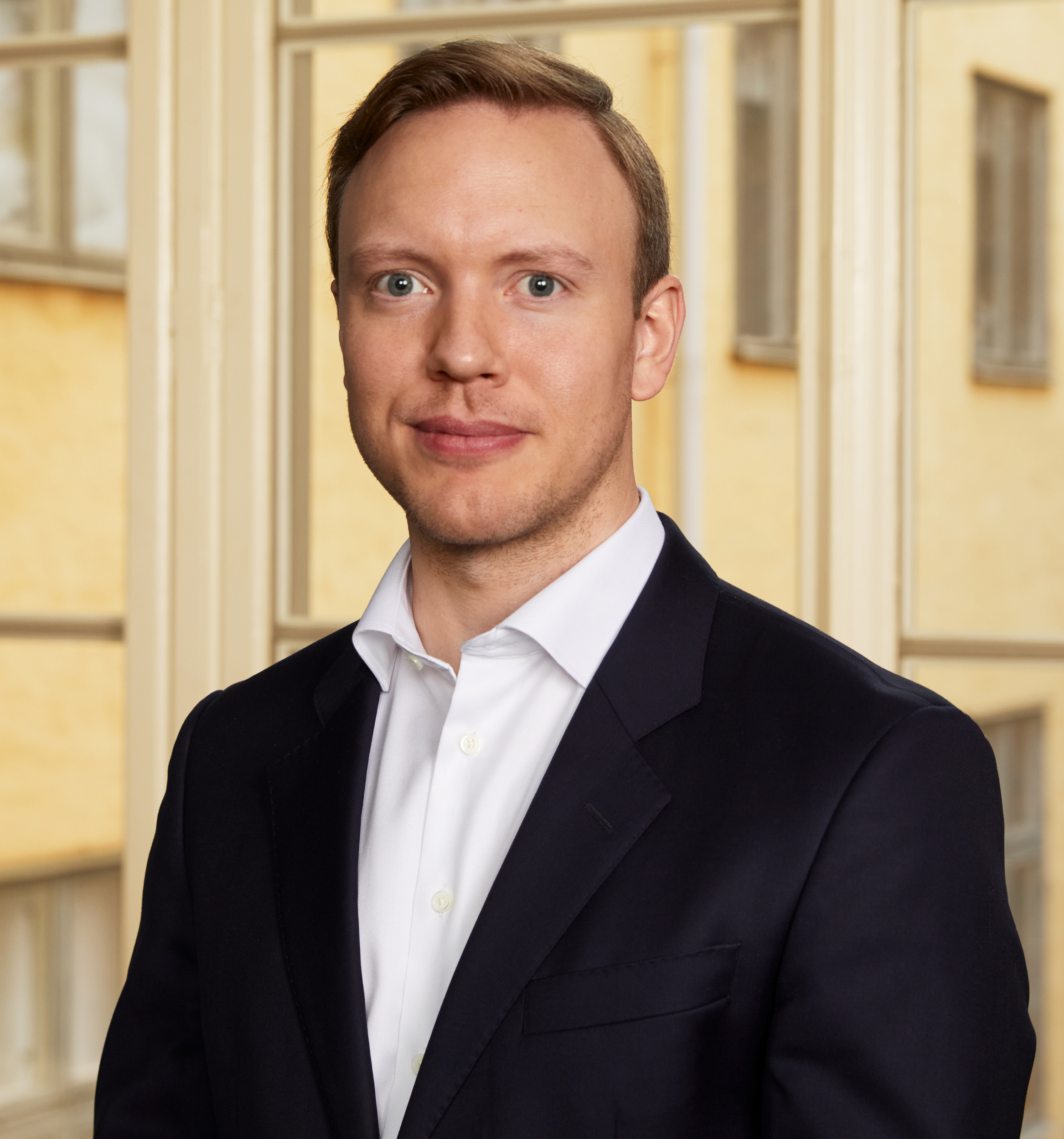 Person in suit with short blonde hair in front of a blurred background of buildings.