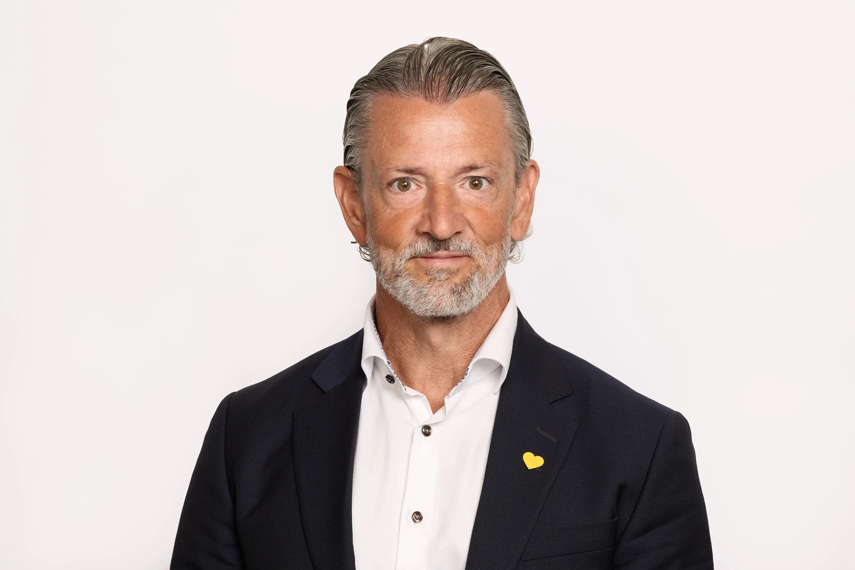 En mann med skjegg i en dress med en gul knapp, foran en hvit bakgrunn.