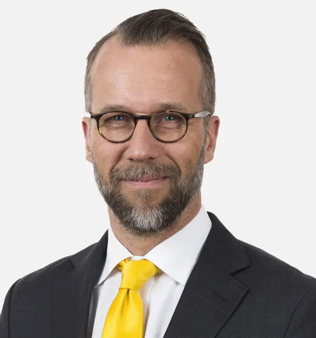 Tom Friberg, press contact - Man in a suit with glasses and a yellow tie, smiling against a white background.