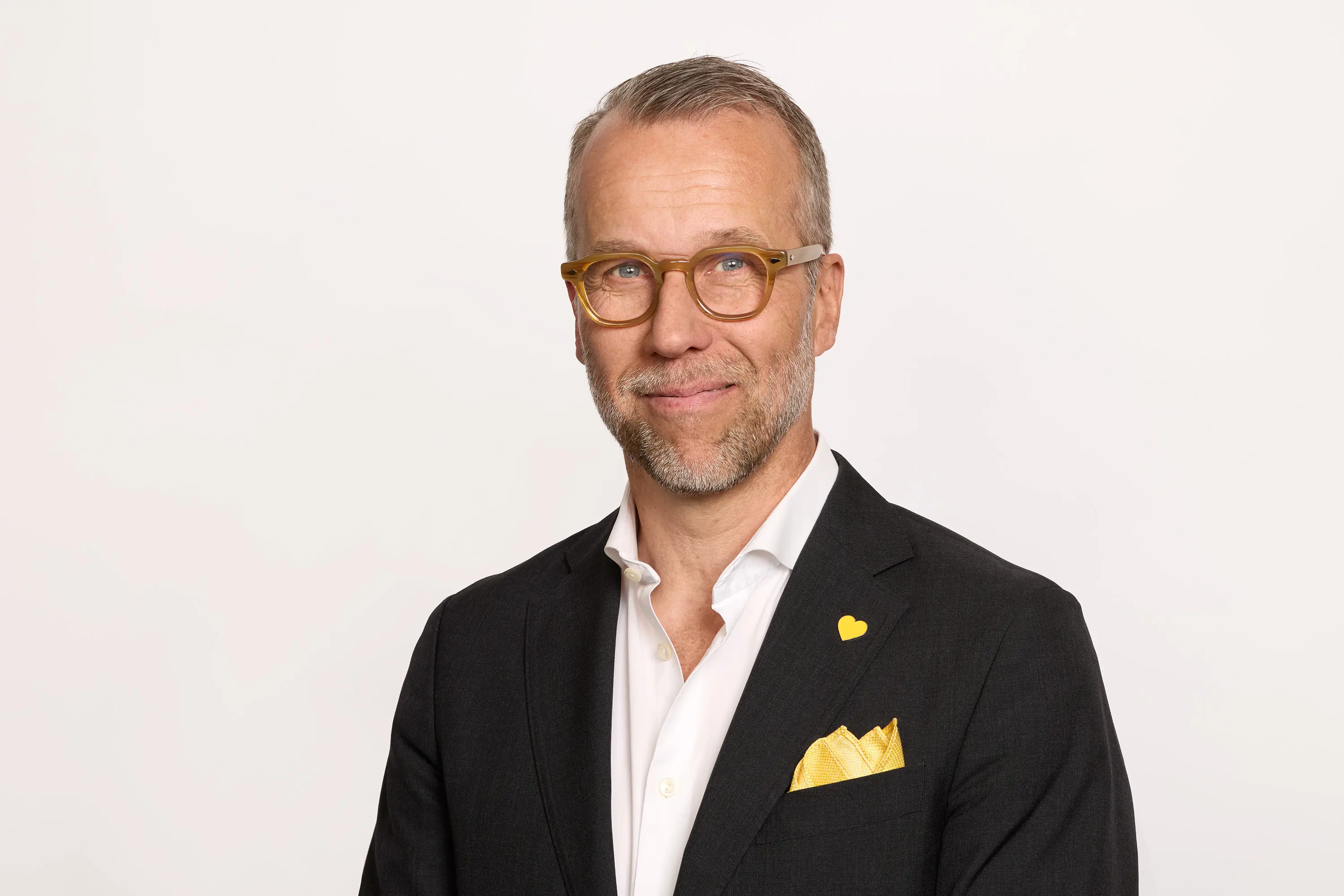 En person med briller i en mørk dress og hvit skjorte står foran en lys bakgrunn. Han har et gult lommetørkle og hjerte på jakkeslaget.