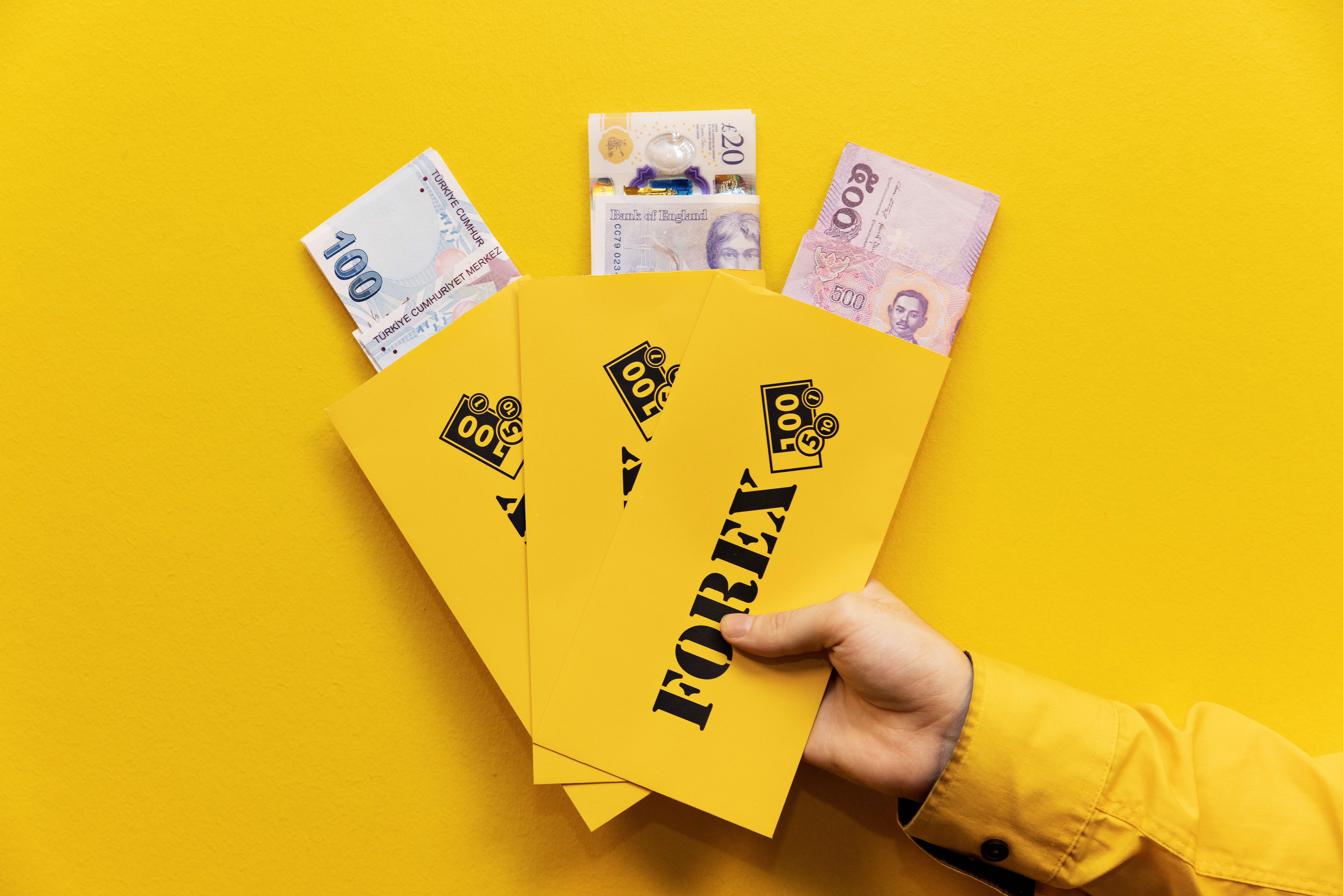 Hand holding Forex envelopes filled with different currencies against yellow background.