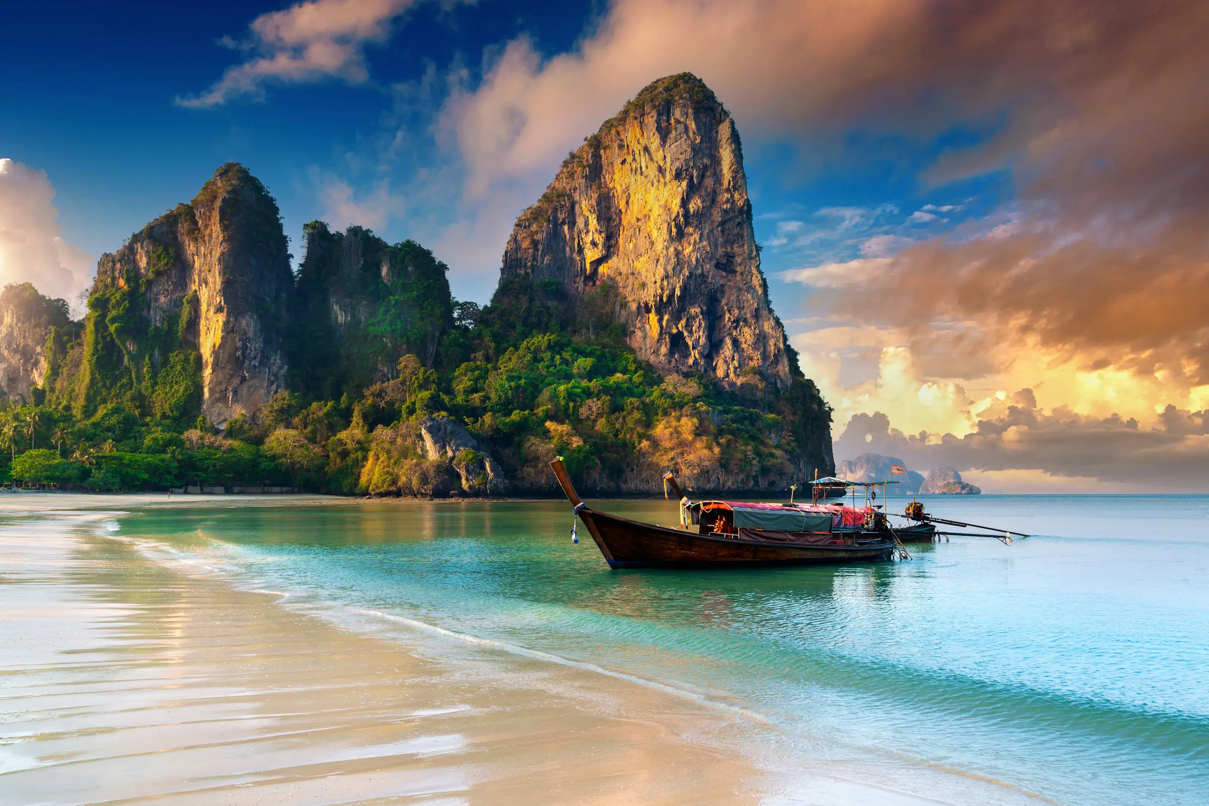Tidlig skumring på en strand som møter blått hav og en longtailbåt med fjell i bakgrunnen i Thailand